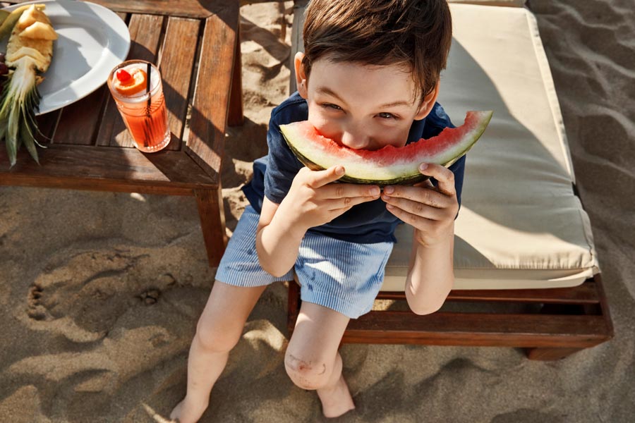 Pojke sitter på en solstol på stranden och äter vattenmelon